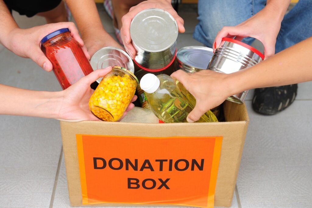 A group of hands placing cans, jars, and bottles of food products into a cardboard donation box.